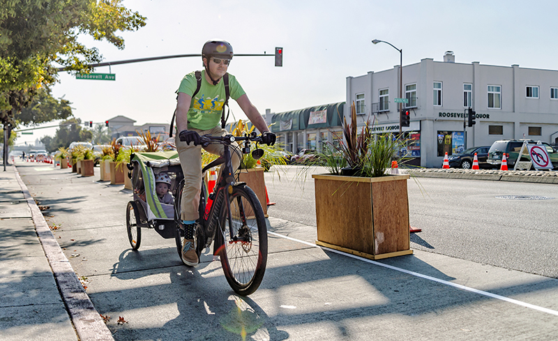 Bicyclist with Kids - Page - Jan2025 Person riding down protected bike lane with child in bike trailer