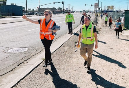 Walking Audit - Page - Jan2025 Group of people in safety vests doing a walking audit along a dirt path to the side of a roadway
