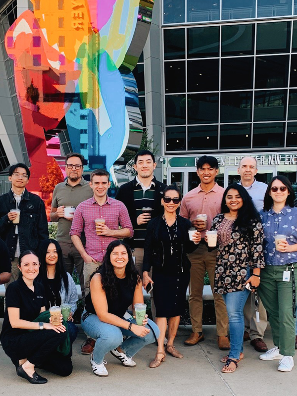 2025 Promotions - blog - July2025 Group of staff gathered in front of colorful art structure holding coffees